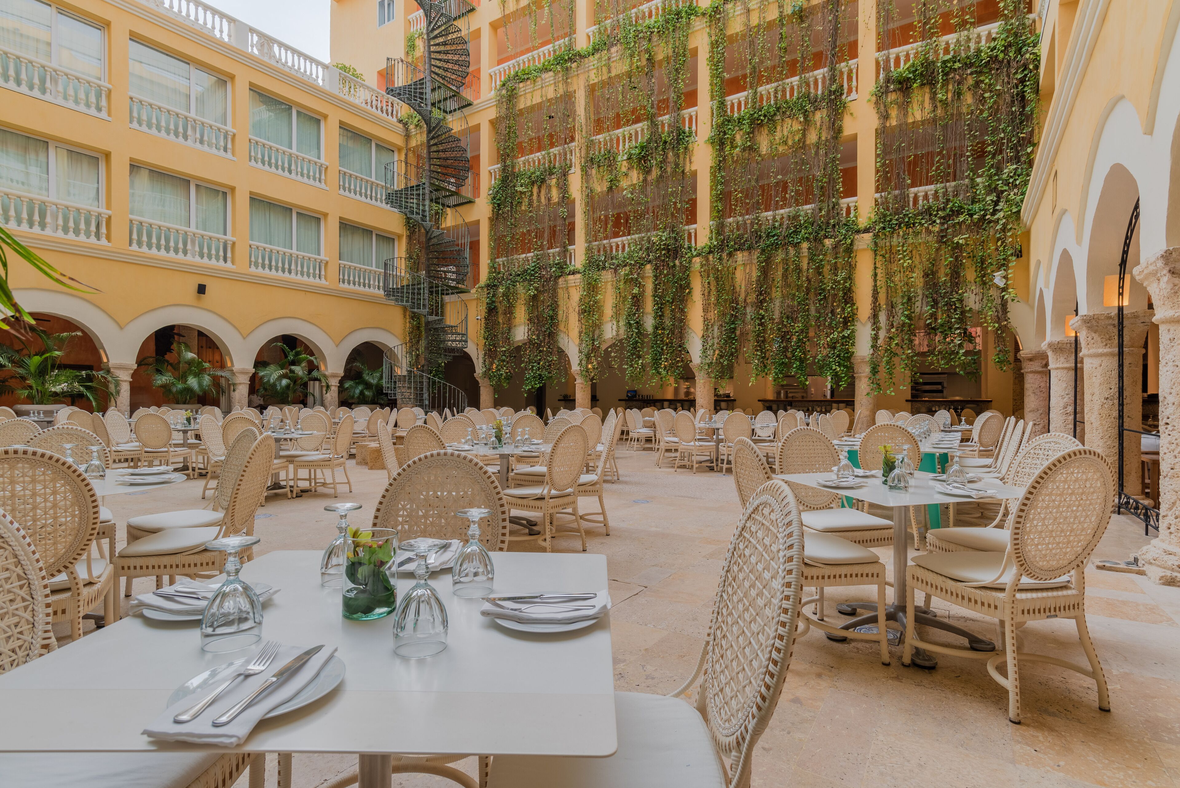 A spacious outdoor dining area with white tables and wicker chairs, surrounded by a yellow building with multiple balconies and a spiral staircase.