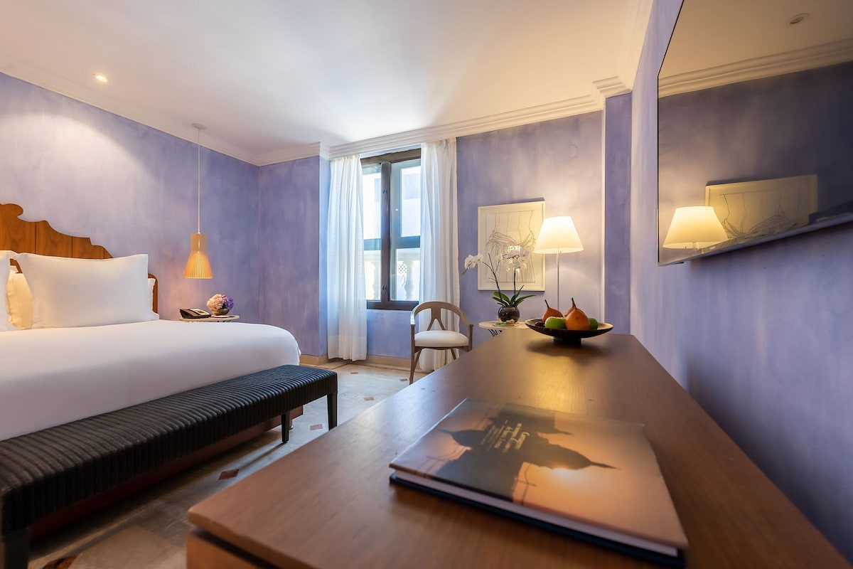 Una habitación de hotel con cama, mesita de noche, lámpara, silla y escritorio con una computadora portátil.