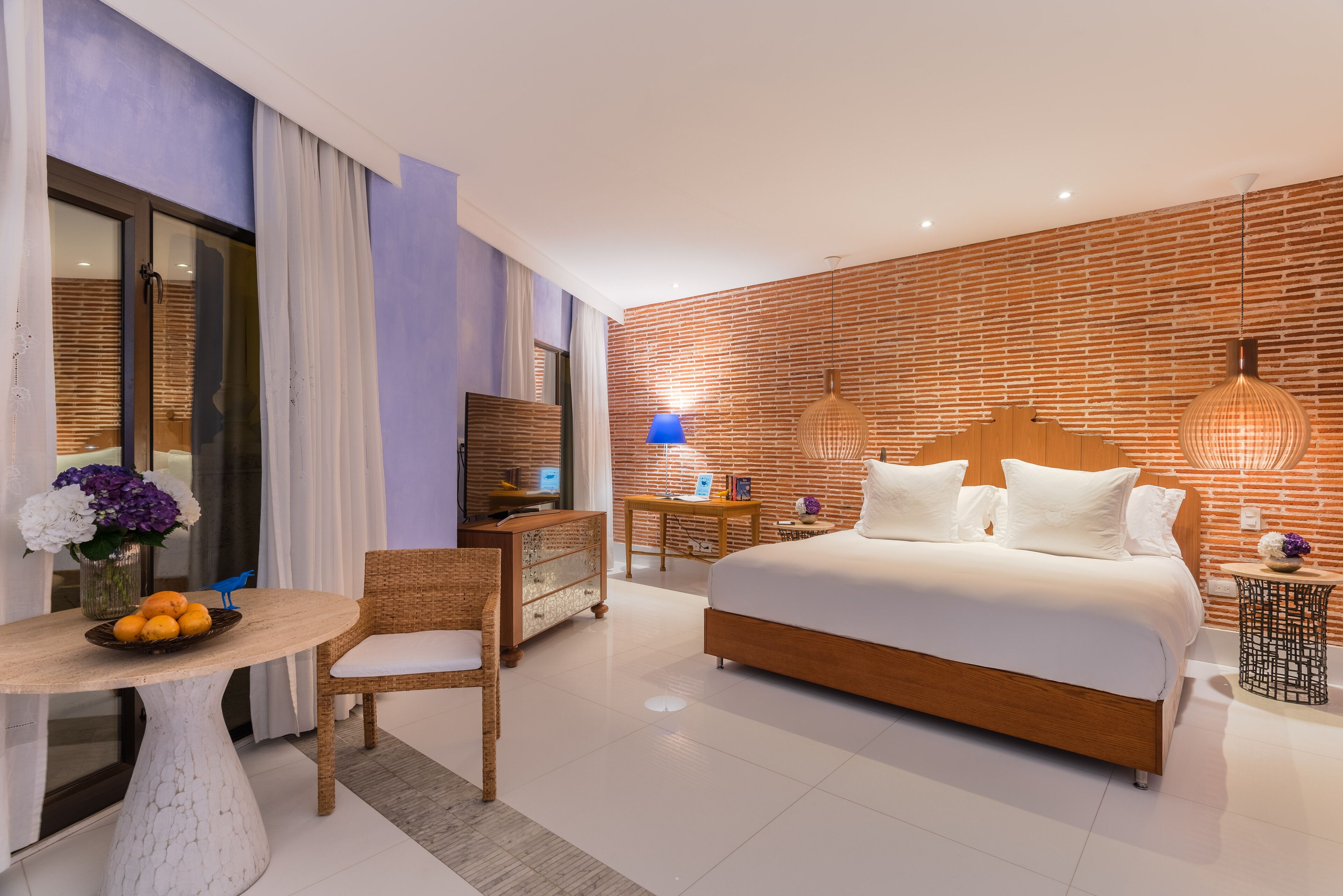 A modern hotel room with a large bed, a desk with a computer, a chair, and a small table with a vase of flowers.