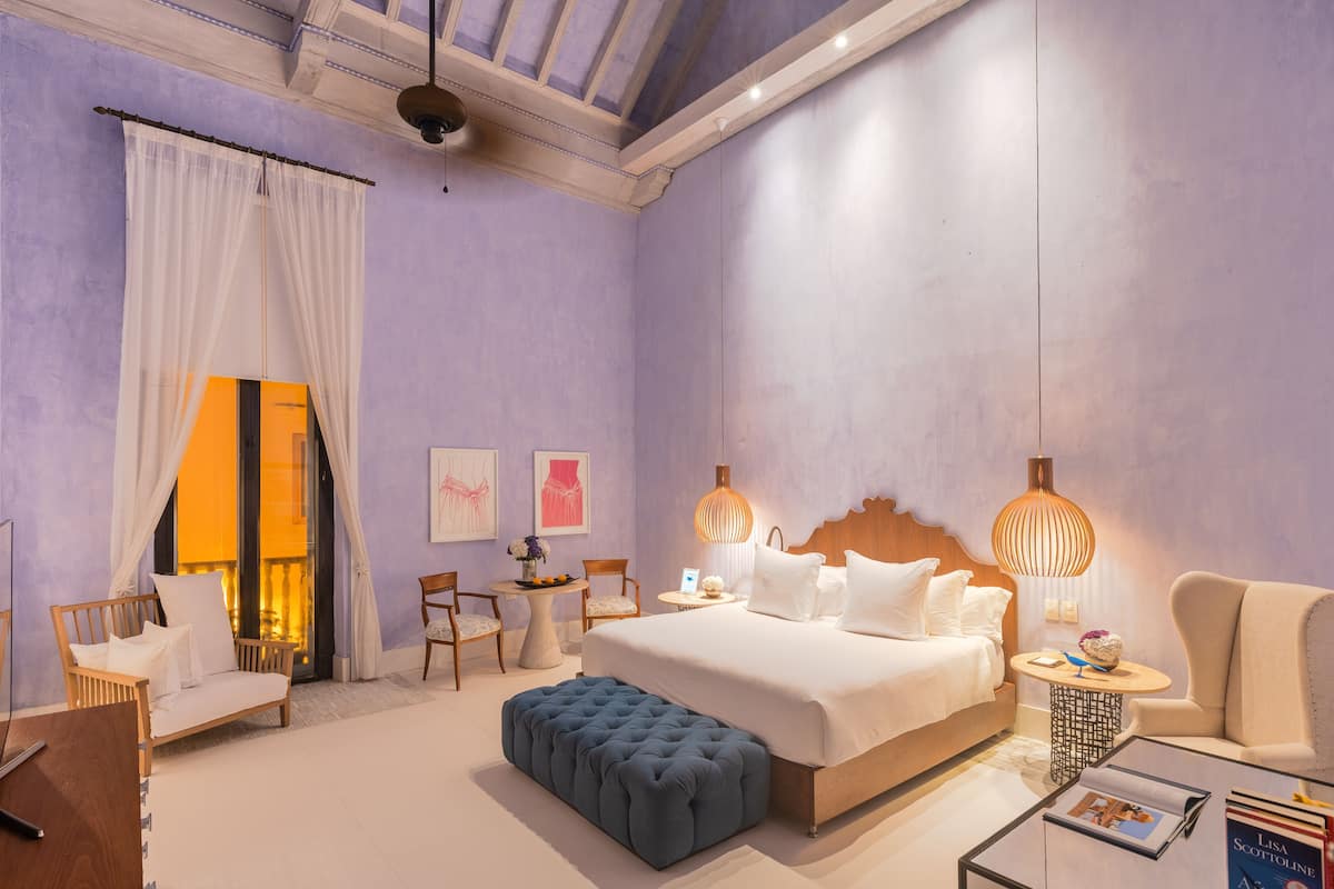 Een slaapkamer met een groot bed, een houten hoofdbord, een blauwe poef, een rond bijzettafeltje en een stoel.