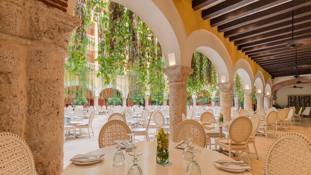 Un vaste espace extérieur aménagé pour les repas, avec des tables blanches et des chaises en osier, entouré d’arcades et de verdure.