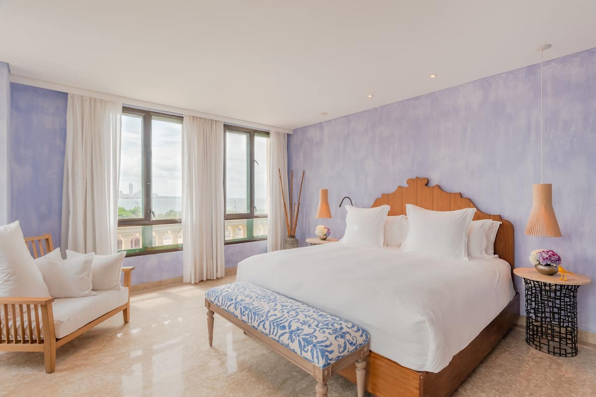 Een hotelkamer met een groot bed, een houten bankje, een rond bijzettafeltje en uitzicht op de stad.