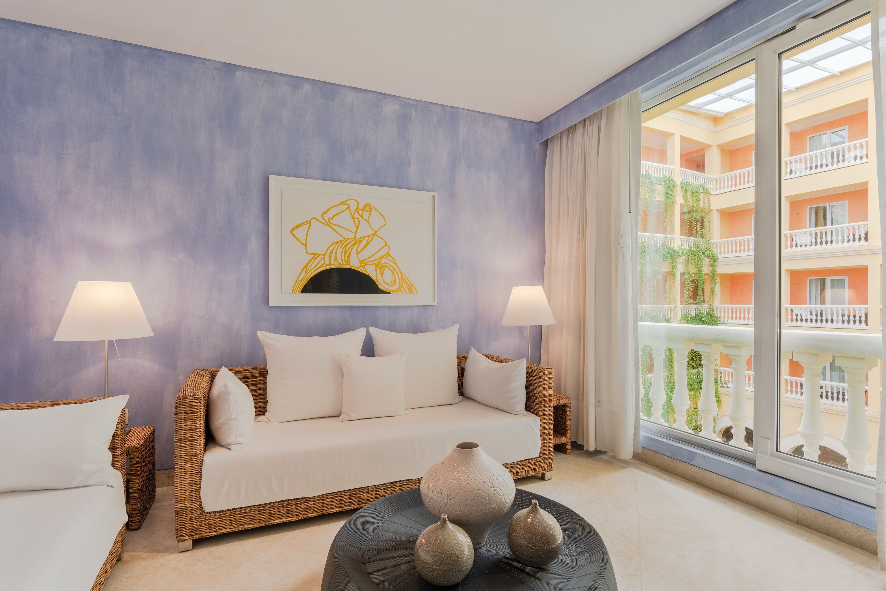 A room with a wicker sofa, two bedside lamps, a vase on a round table, and a large window with a view of a building.