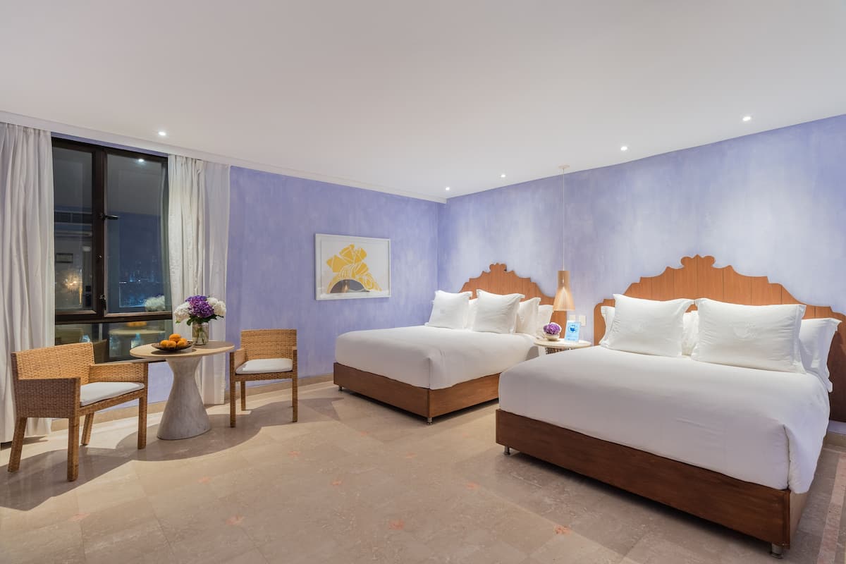Een hotelkamer met twee bedden, een tafeltje met stoelen, een raam met gordijnen en uitzicht op de stad.