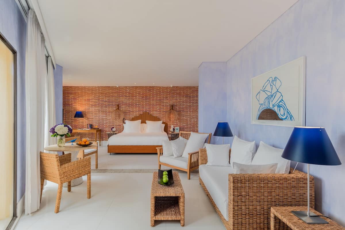 Een moderne hotelkamer met een groot bed, een zithoek met rieten meubels, een eettafel met een bloevas en een blauwe lamp.