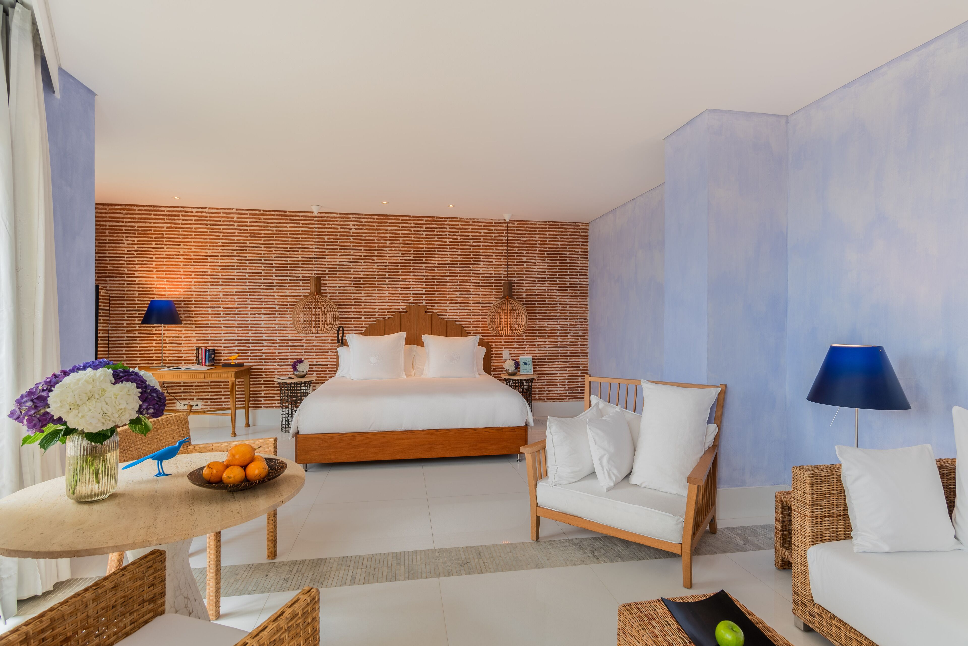 Una habitación de hotel con una cama grande, un cabecero de madera, una mesa de comedor con un jarrón de flores, una silla de mimbre y una lámpara azul.