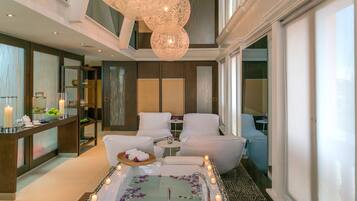 Un área de spa con una bañera grande rodeada de velas, dos sillones blancos y una mesita con un frutero y flores.