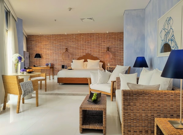 Una habitación de hotel moderna con pared de ladrillo, una cama grande, una zona de comedor con mesa y sillas, y un espacio de descanso con mobiliario de mimbre.