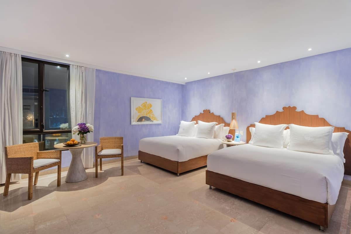 Een hotelkamer met twee bedden, een klein tafeltje met een stoel, een raam met gordijnen en een wanddecoratie.