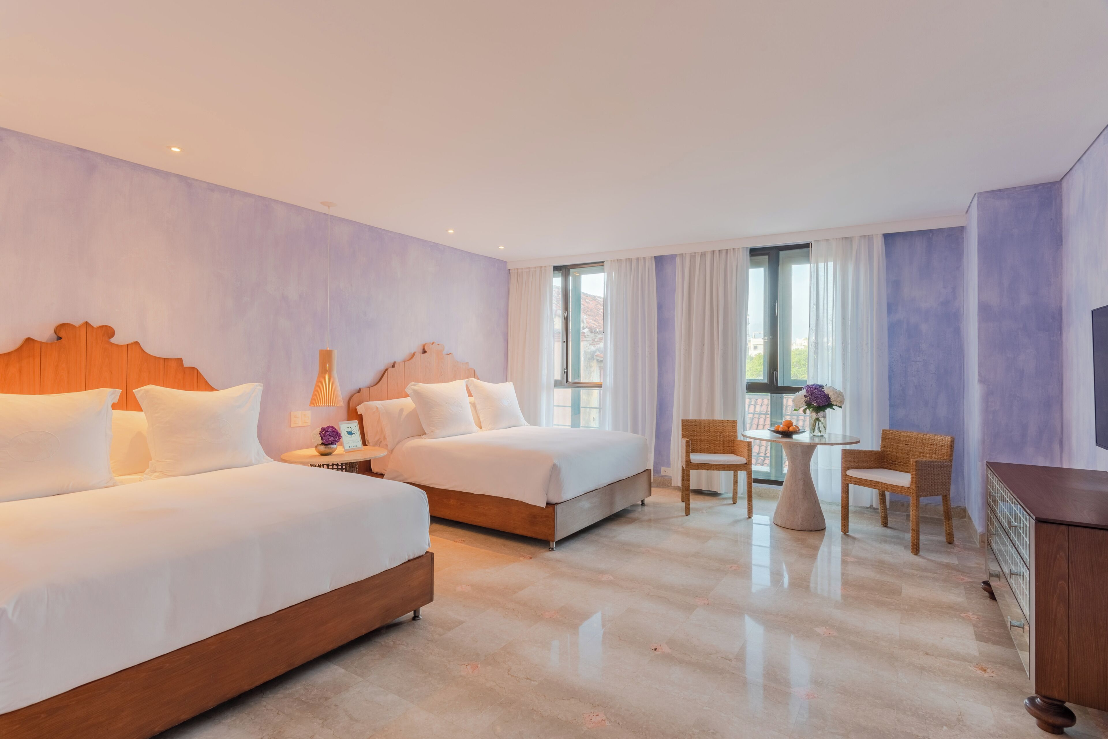 A hotel room with two beds, a dining table with chairs, a TV, and a window with curtains.