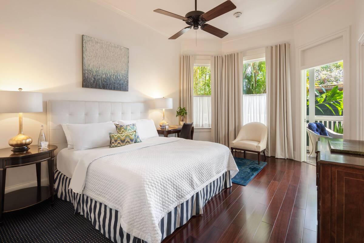 A hotel room with a large bed, a ceiling fan, a window with curtains, a desk with a lamp, and a view of greenery outside.