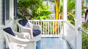 A covered porch with white wicker furniture and a blue floor, surrounded by lush tropical plants.