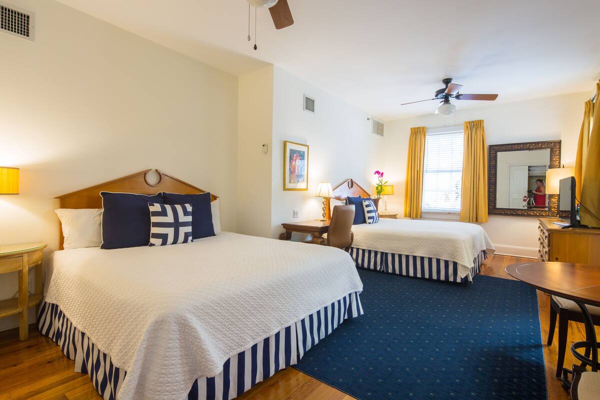 A hotel room with two beds, a desk, a chair, a ceiling fan, and a mirror.