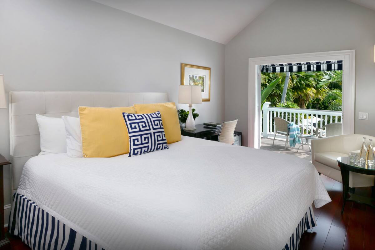 A neatly made bed with white and yellow pillows, a bedside table with a lamp, and a view of a balcony with outdoor furniture.
