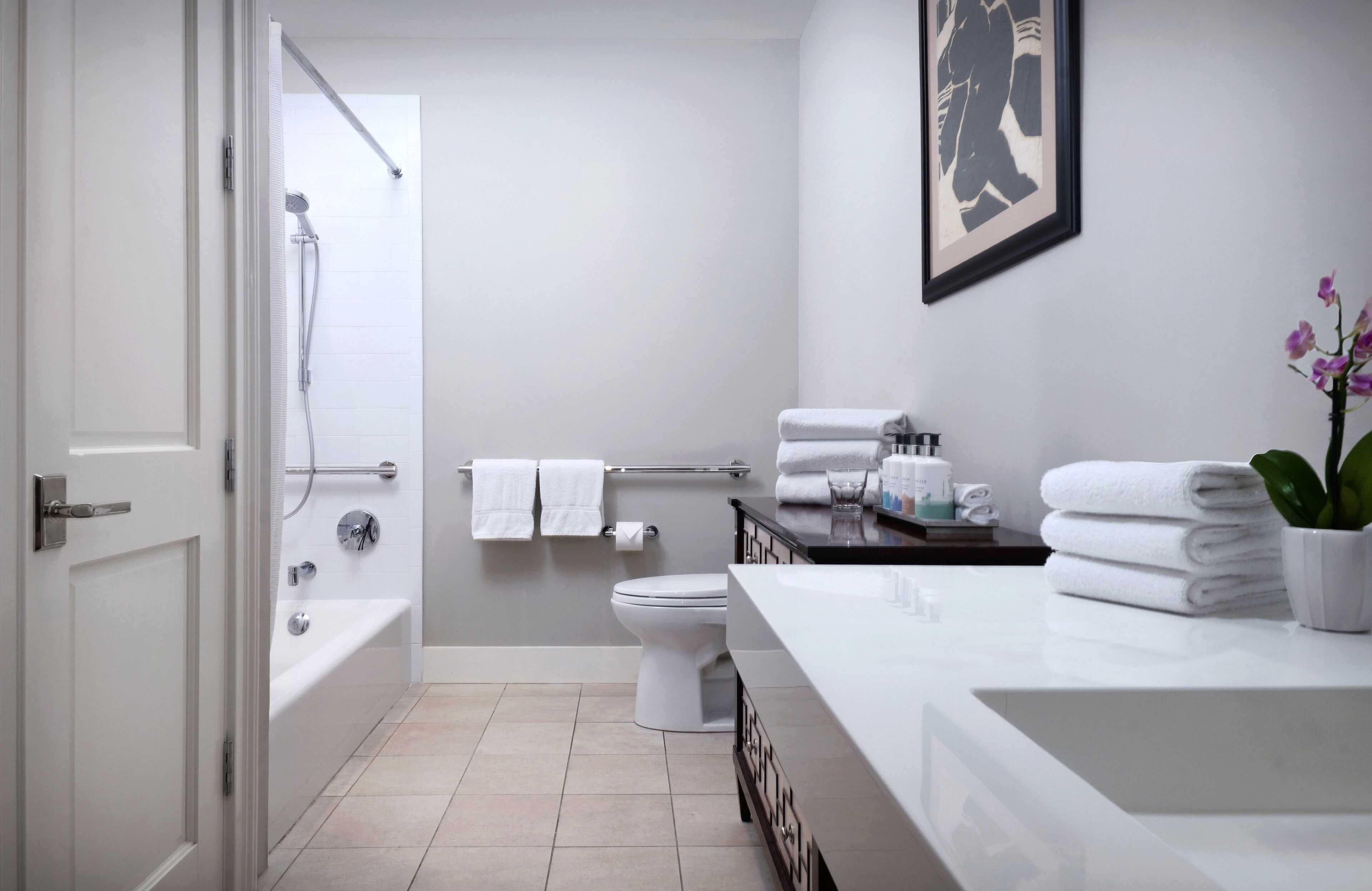 A modern bathroom with a bathtub, toilet, and sink. There are towels stacked on the counter and a framed picture on the wall.