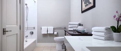 A modern bathroom with a bathtub, toilet, and sink. There are towels stacked on the counter and a framed picture on the wall.