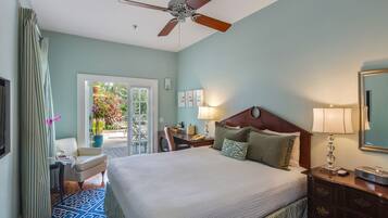 A hotel room with a bed, a desk, a chair, a ceiling fan, a nightstand, and a window with a view of plants and a patio.