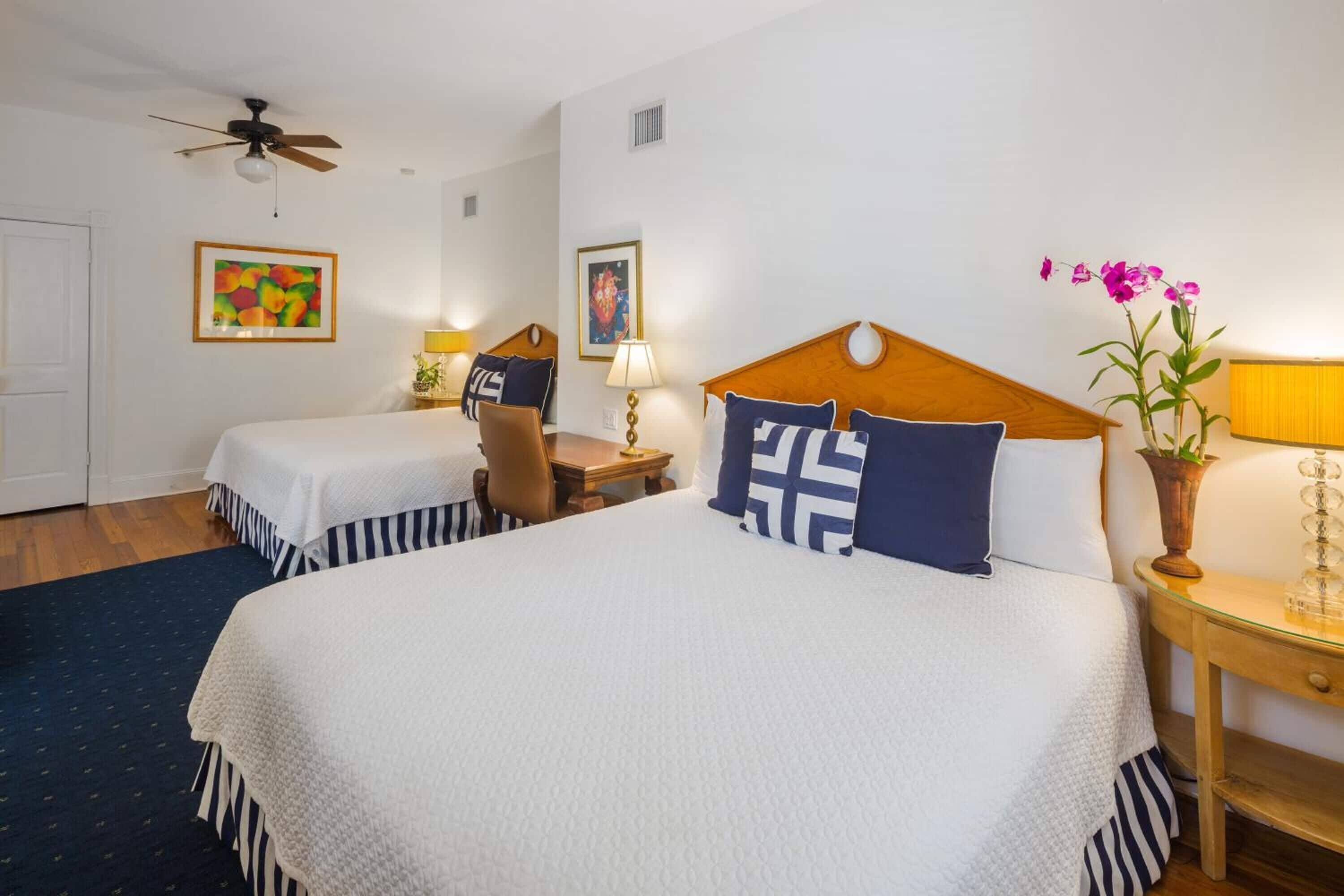 A hotel room with a large bed, two smaller beds, a desk, and a ceiling fan.
