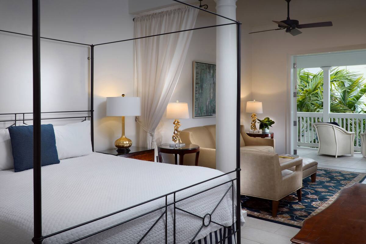 A four-poster bed with a white bedspread and blue pillow, a round table with a lamp, a beige armchair, and a view of a balcony with palm trees.