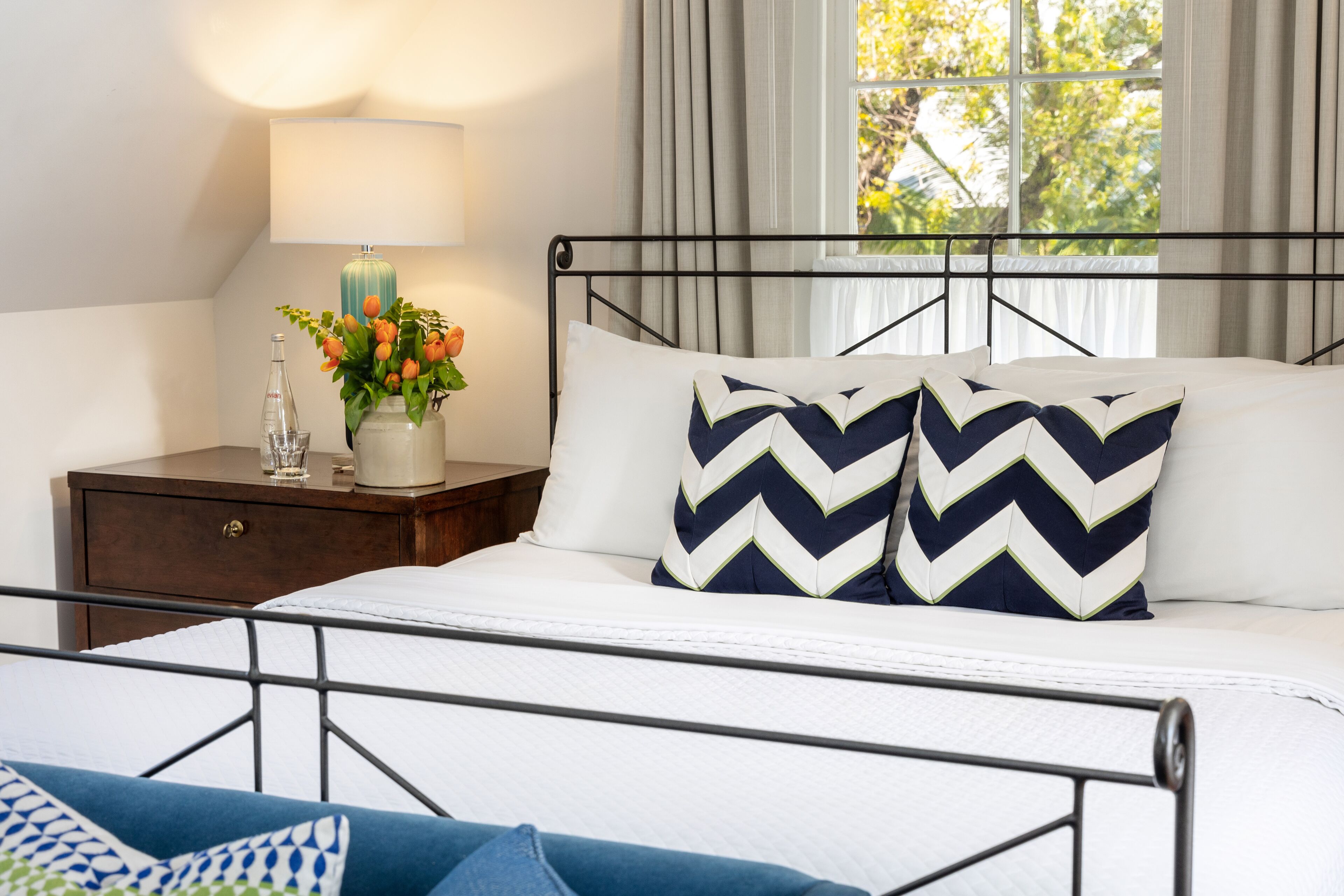 Una cama bien hecha con estructura metálica, una mesita de noche con una lámpara y un florero, y una ventana con cortinas.