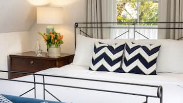 Una cama bien hecha con estructura metálica, una mesita de noche con una lámpara y un florero, y una ventana con cortinas.