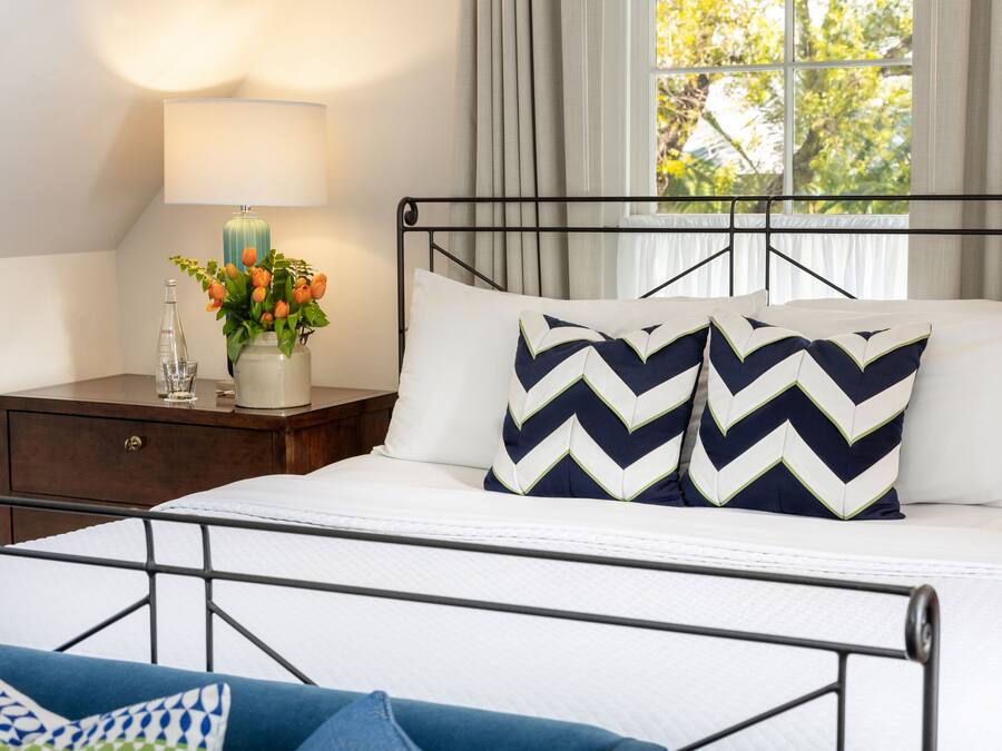 Una cama bien hecha con estructura metálica, una mesita de noche con una lámpara y un florero, y una ventana con cortinas.
