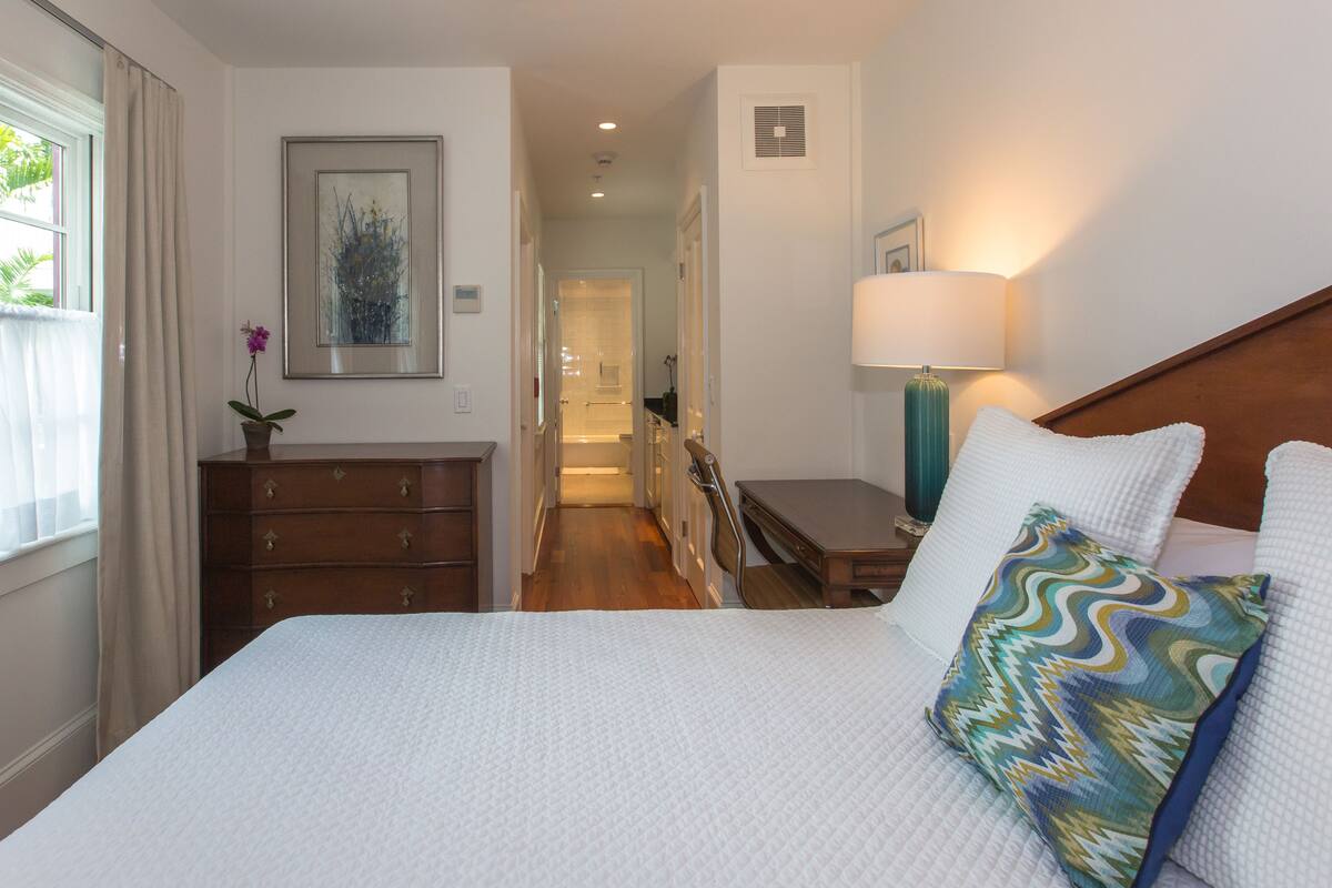 A hotel room with a bed, a dresser, a nightstand, a lamp, and a bathroom visible through an open door.