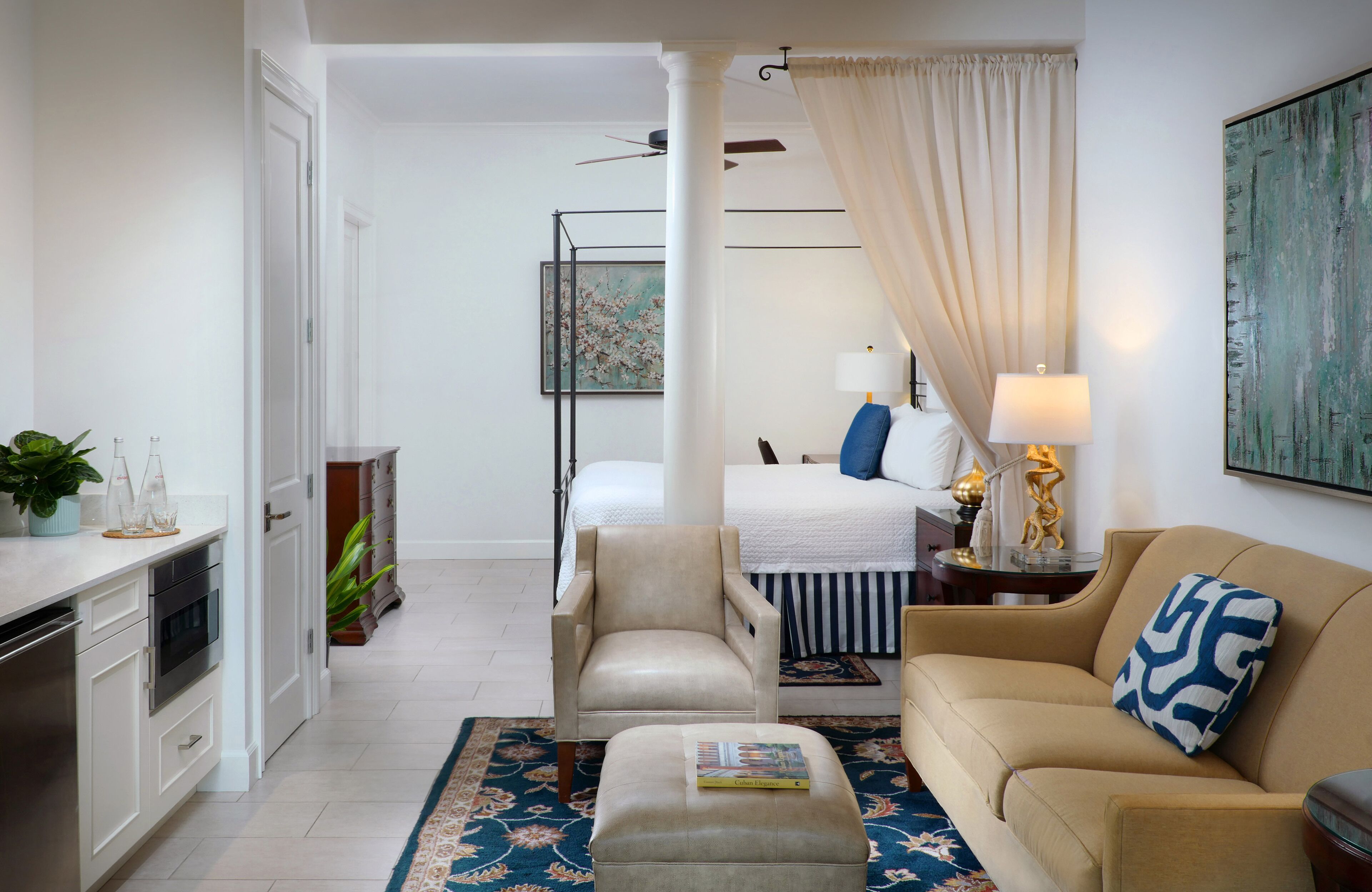 A modern living room with a beige sofa, a matching ottoman, and a coffee table. There is a bed with a striped bedspread and a nightstand with a lamp. A large painting hangs on the wall, and a window with curtains is visible.