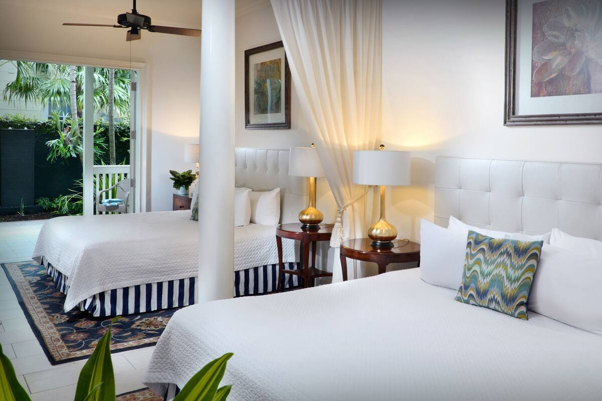 A hotel room with two beds, a ceiling fan, a large window with a view of palm trees, and a painting on the wall.