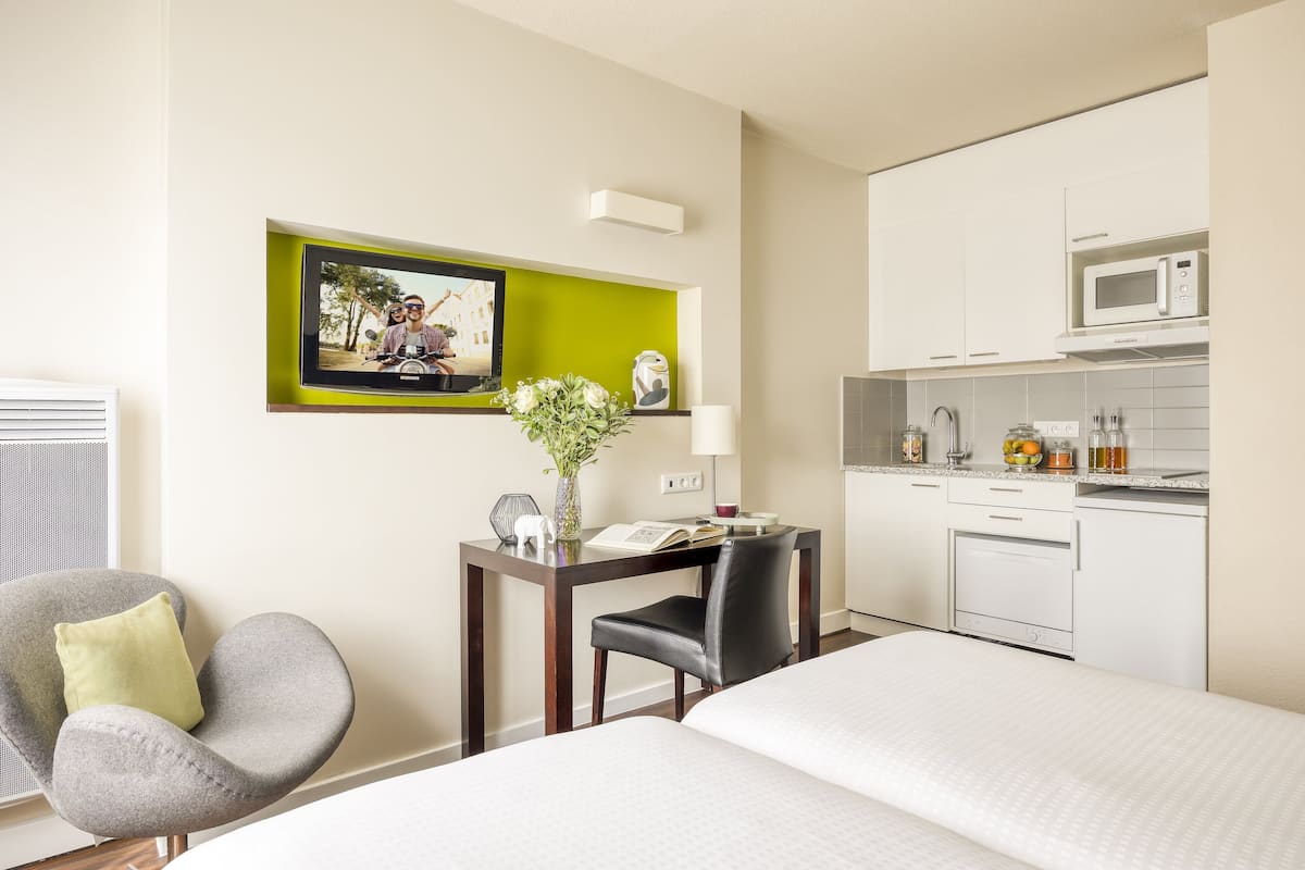 Quarto de hotel com uma cama, uma escrivaninha com cadeira, micro-ondas e uma pequena cozinha equipada.