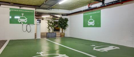 Area di parcheggio interna con pavimento verde, linee di parcheggio bianche e segnaletica per punti di ricarica veicoli elettrici.