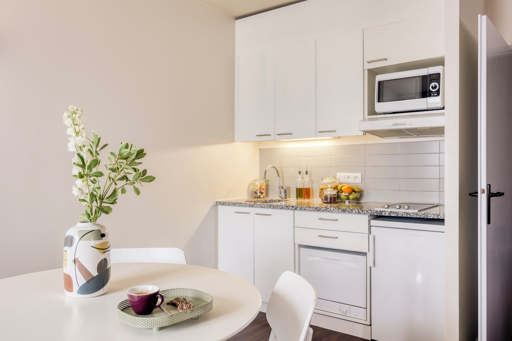 A modern kitchen with white cabinets, a microwave, and a dining area with a table, chairs, and a vase with flowers.