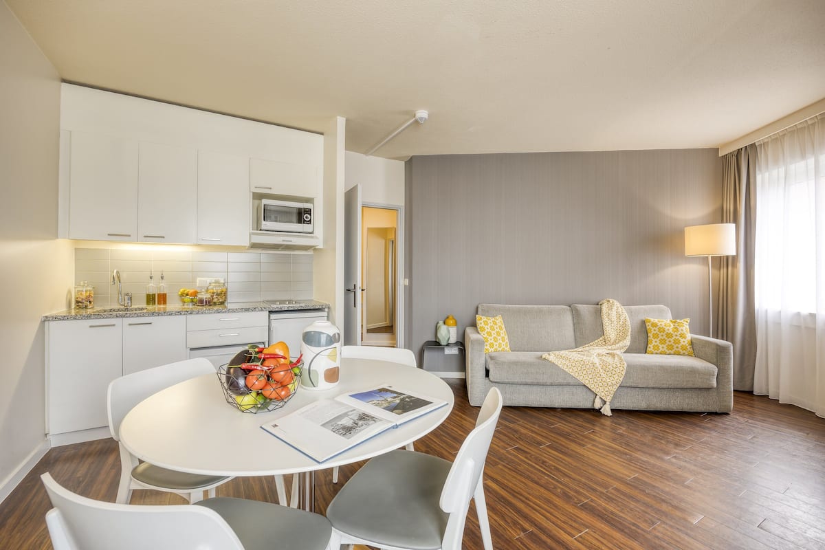 Uma sala de estar moderna com um sofá cinzento, uma mesa de jantar com cadeiras e uma área de cozinha com armários brancos e micro-ondas.