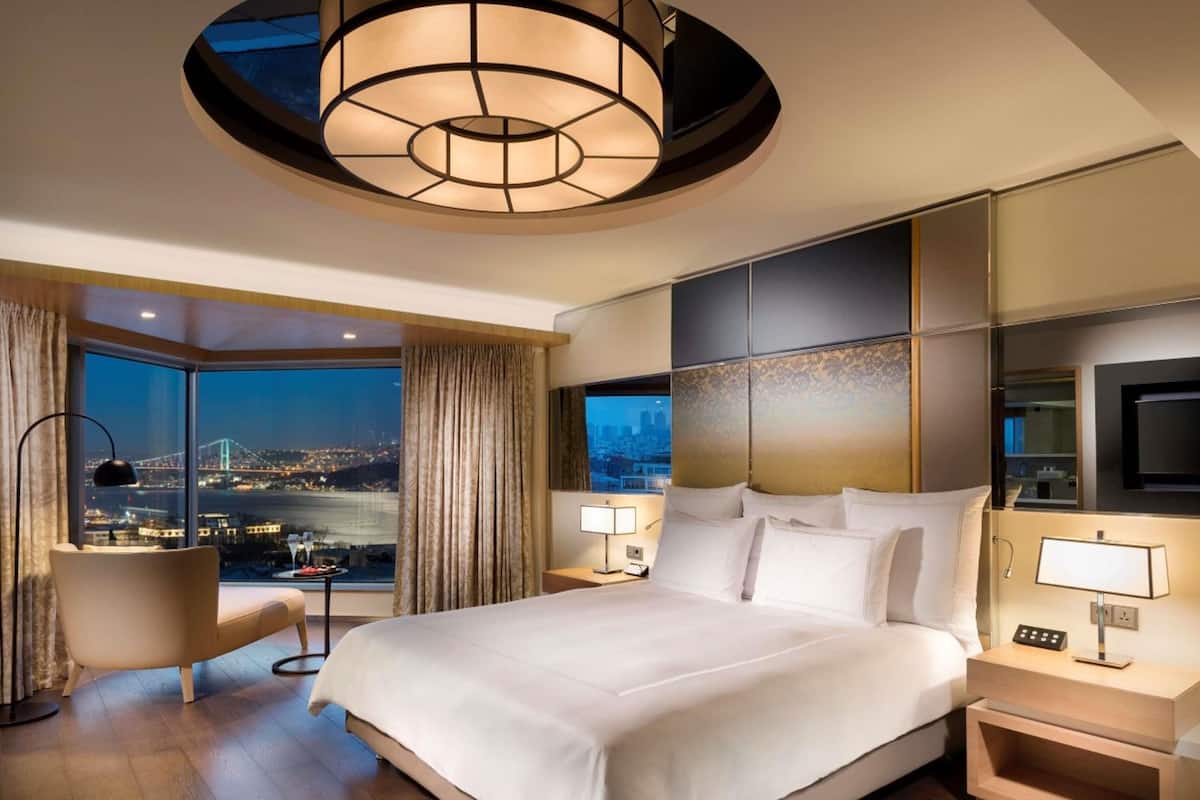 A modern hotel room with a large bed, a desk, a chair, a TV, and a view of a cityscape.