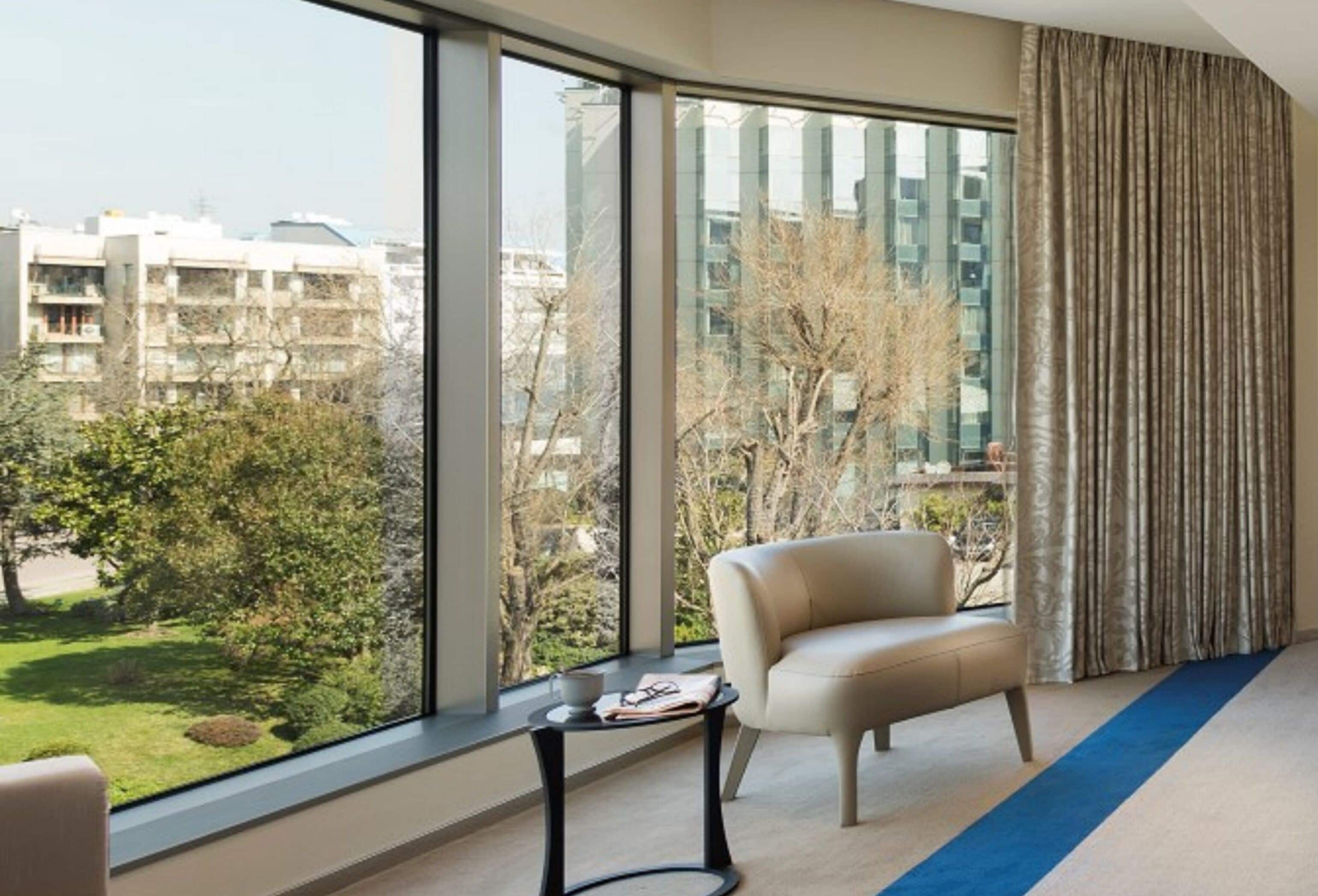 A modern hotel room with a large window, a beige sofa, a small side table, and a blue carpet.