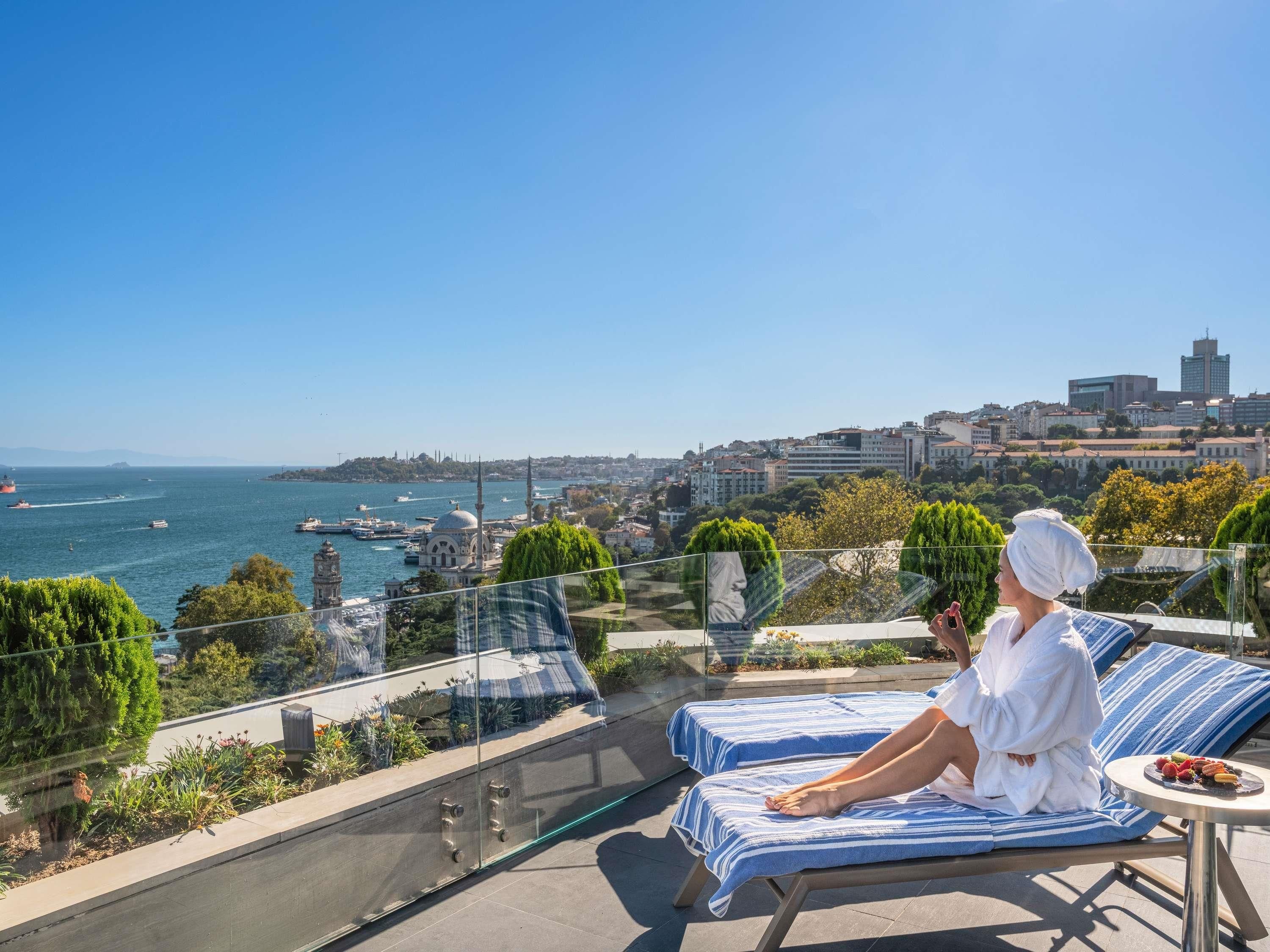 Un balcon avec vue sur la mer et la ville, une personne se relaxant sur un fauteuil et une table avec des fruits.