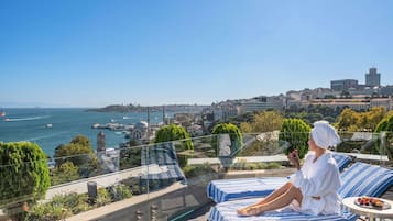 Un balcon avec vue sur la mer et la ville, une personne se relaxant sur un fauteuil et une table avec des fruits.