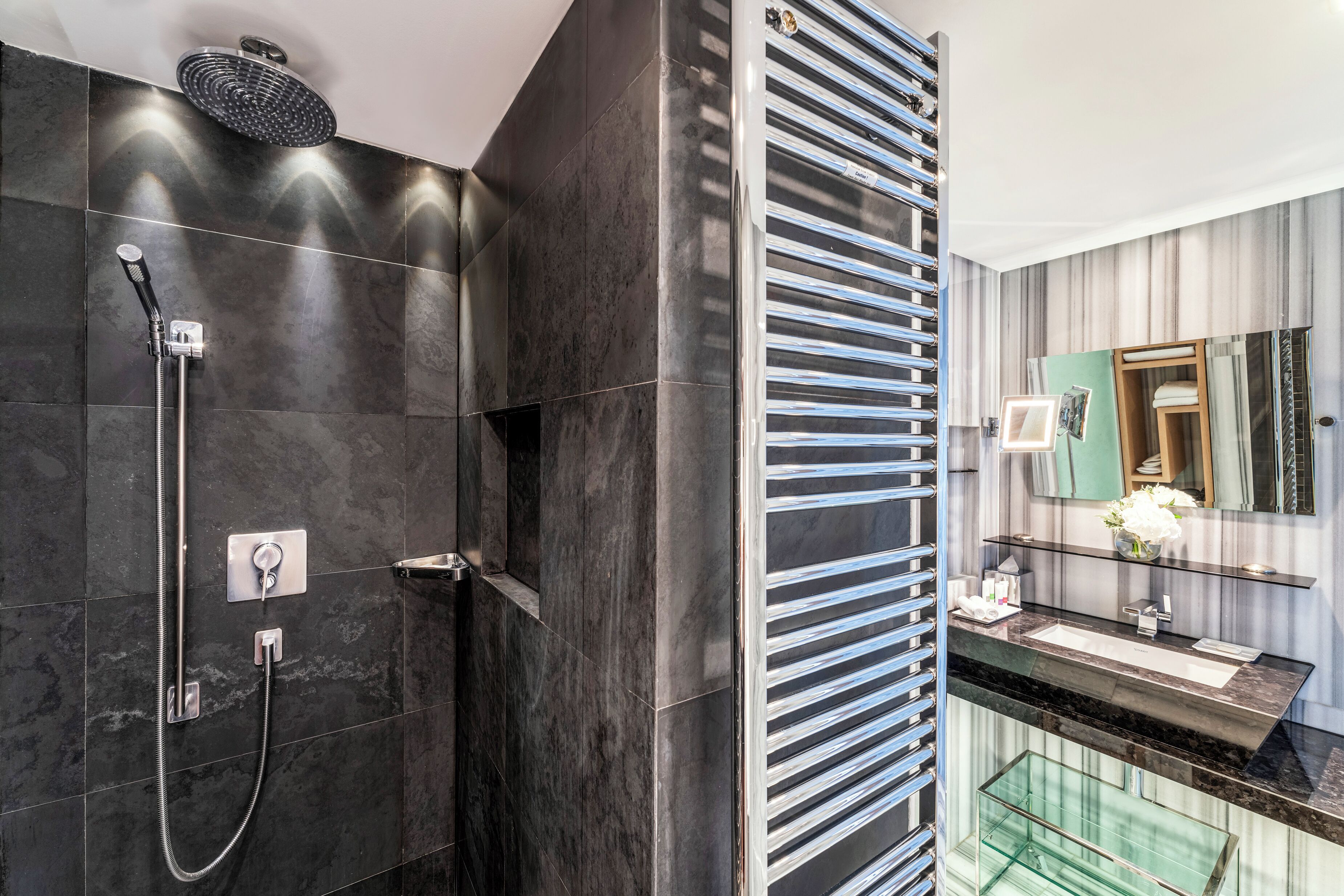 A modern bathroom with a glass shower enclosure, a dark tiled wall, a vanity with a mirror, and a glass shelf.
