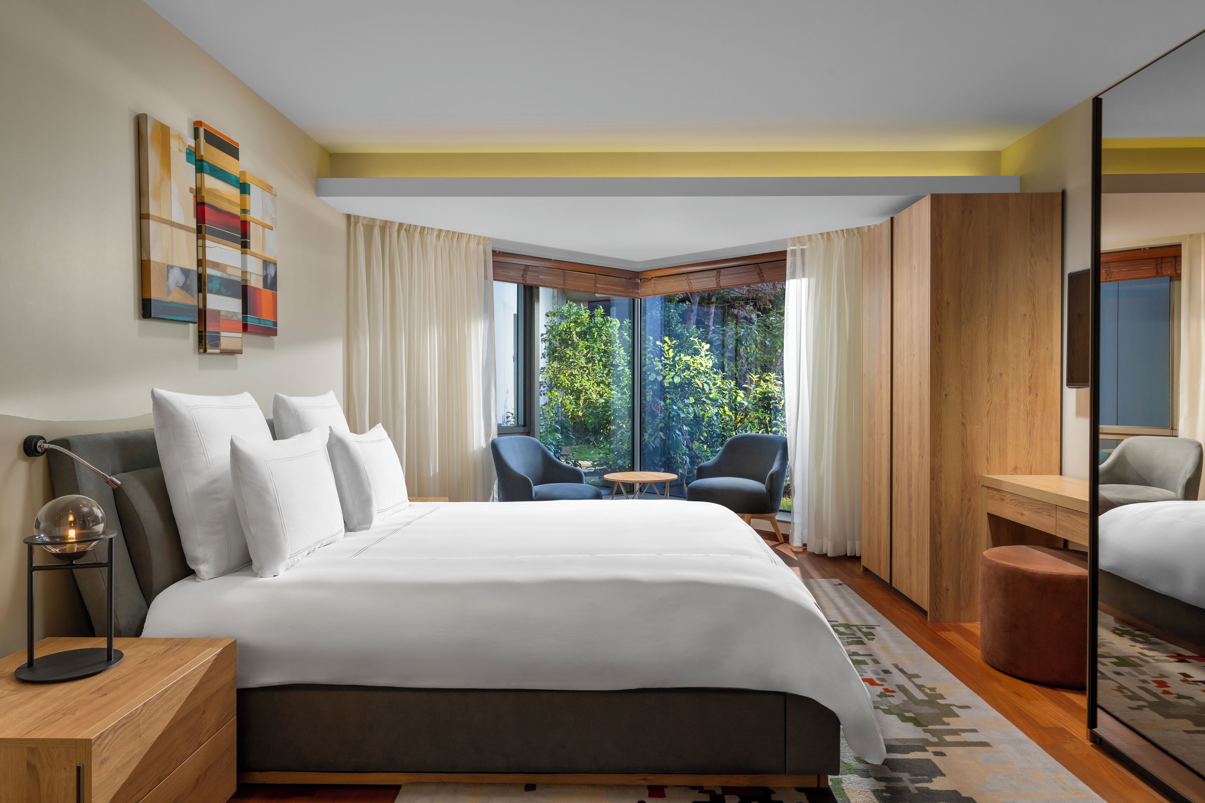 A modern hotel room with a large bed, two armchairs, a small table, and a view of greenery through large windows.