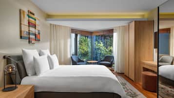 A modern hotel room with a large bed, two armchairs, a small table, and a view of greenery through large windows.