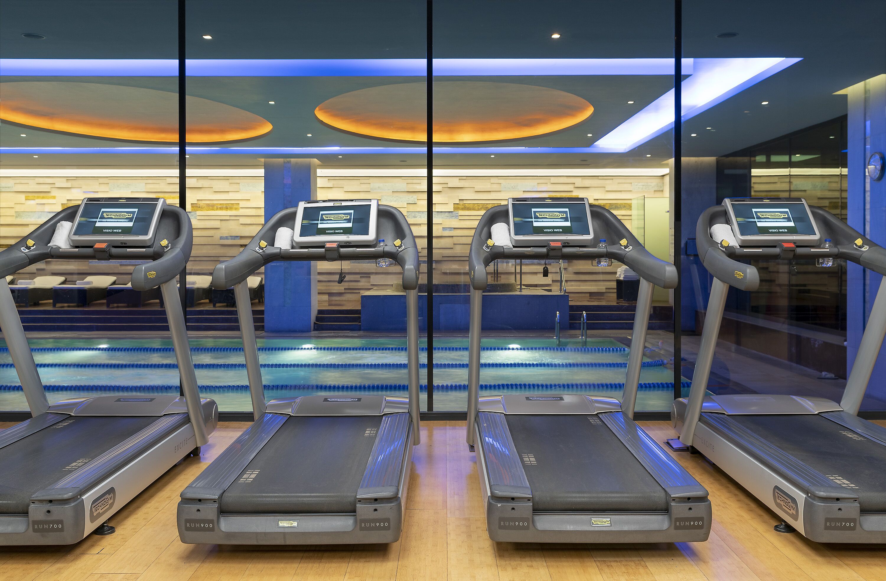 Cuatro cintas de correr con pantallas digitales en un gimnasio con piscina al fondo.