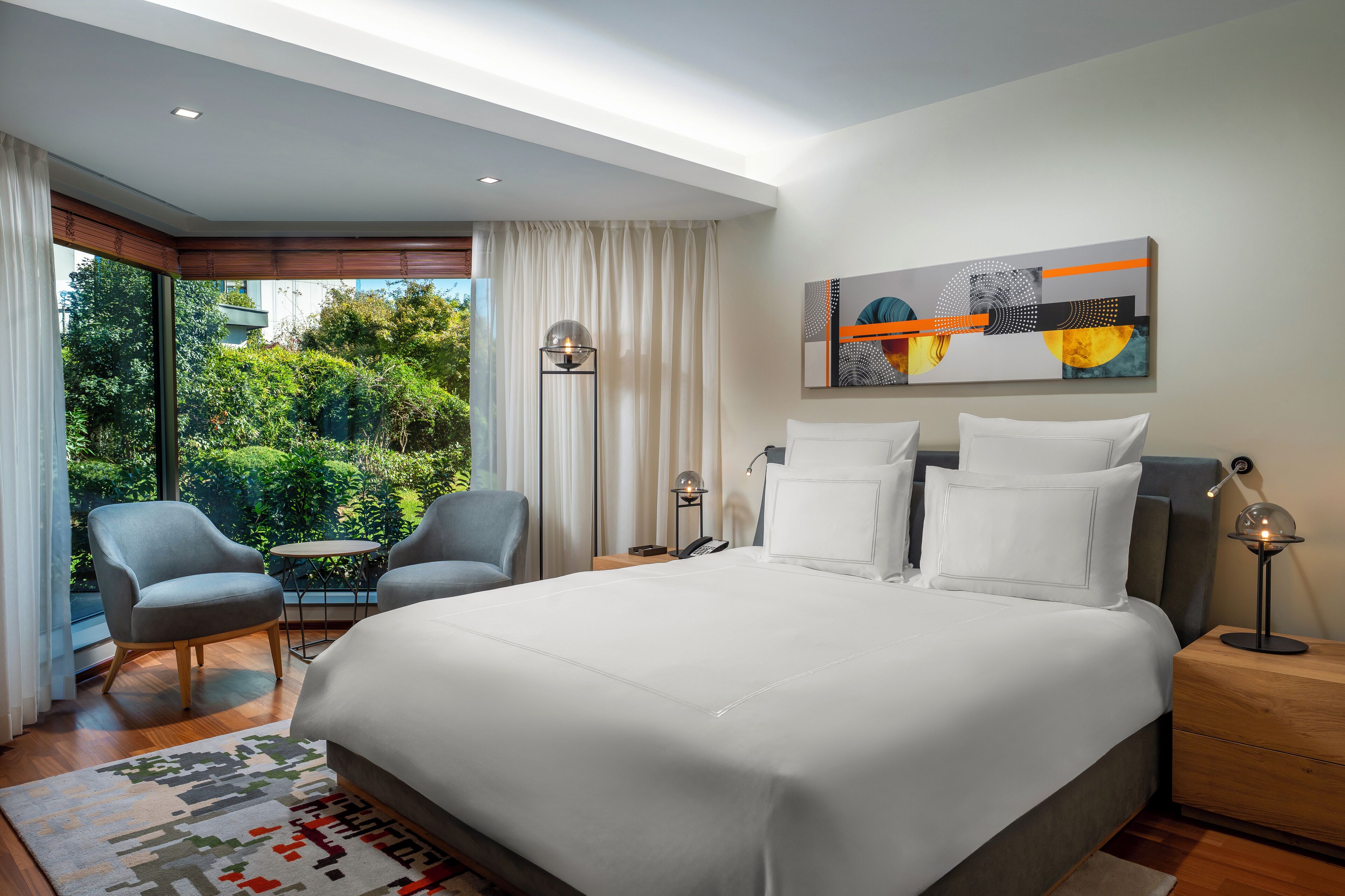 A modern hotel room with a large bed, two armchairs, a small table, and a view of greenery through large windows.