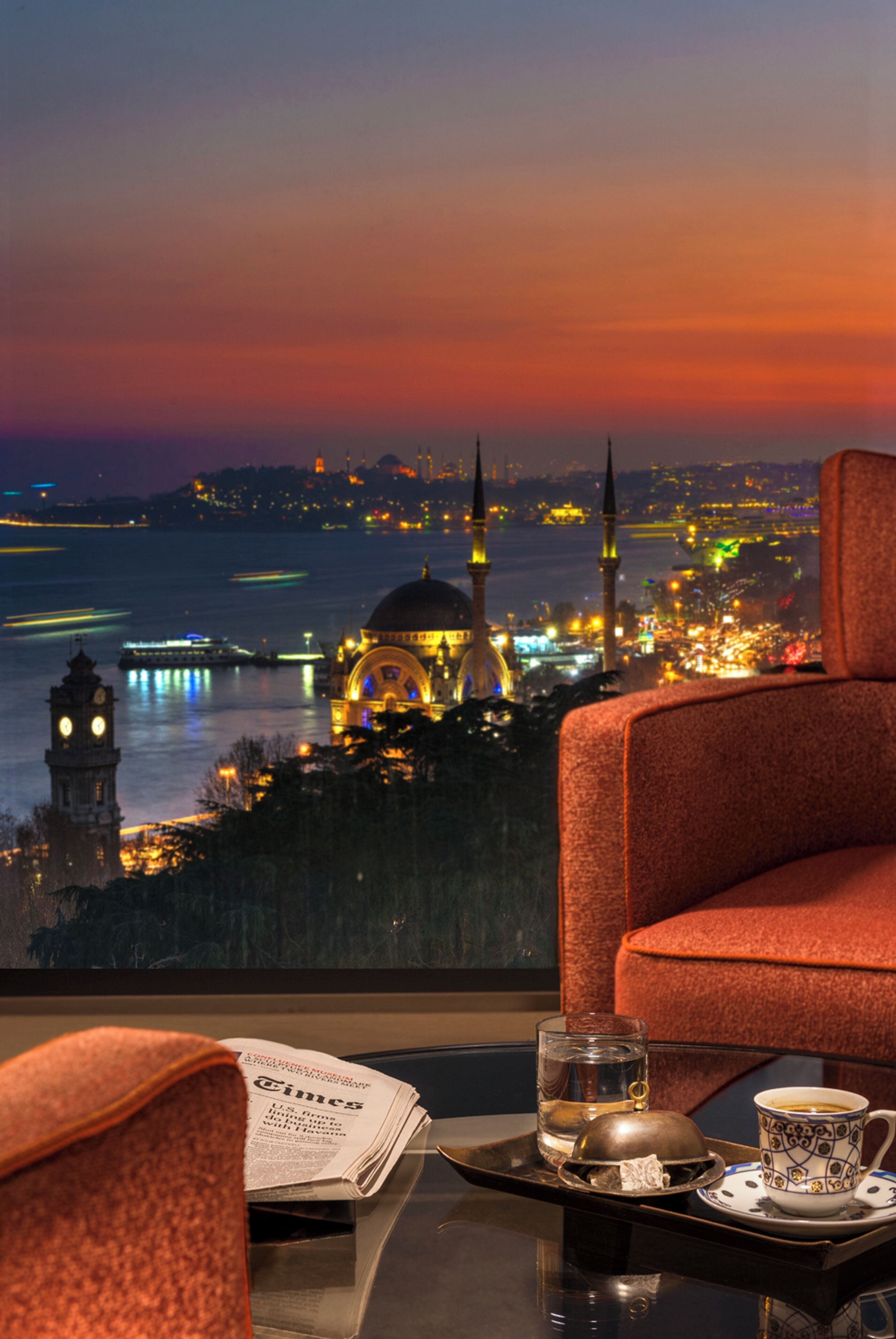 Habitación de hotel con vista a un paisaje urbano al atardecer, que incluye una torre del reloj y una mezquita.