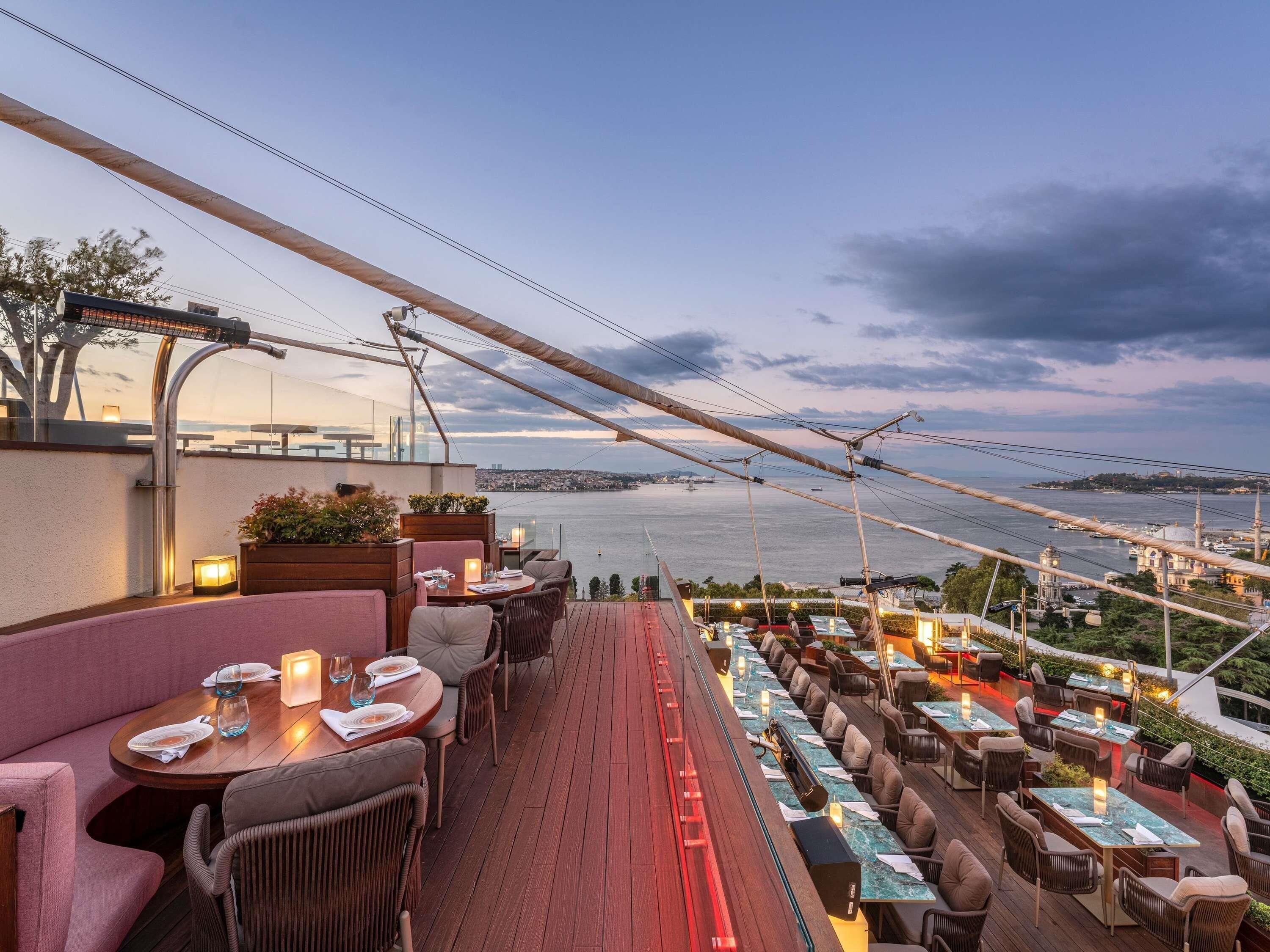 An outdoor dining area with tables and chairs, overlooking a body of water and a cityscape.