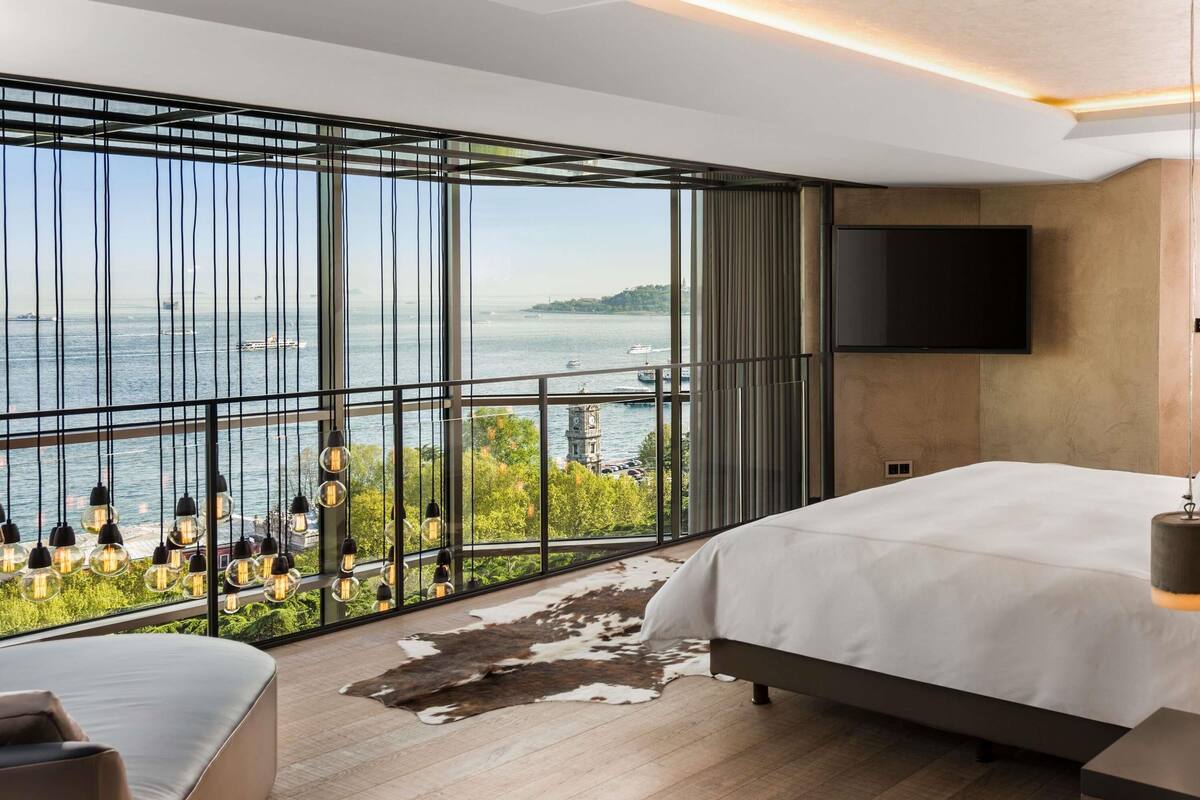 A modern bedroom with a large window, a bed, a TV, and a view of the water.