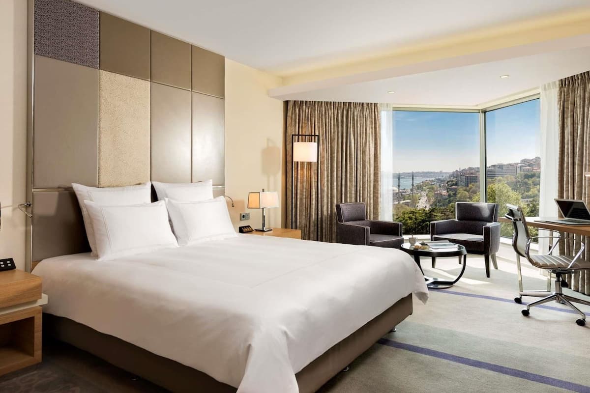 Una habitación de hotel con una cama grande, un escritorio con una computadora portátil, dos sillas, vistas de un paisaje urbano a través de ventanas amplias y una mesita de noche con una lámpara.
