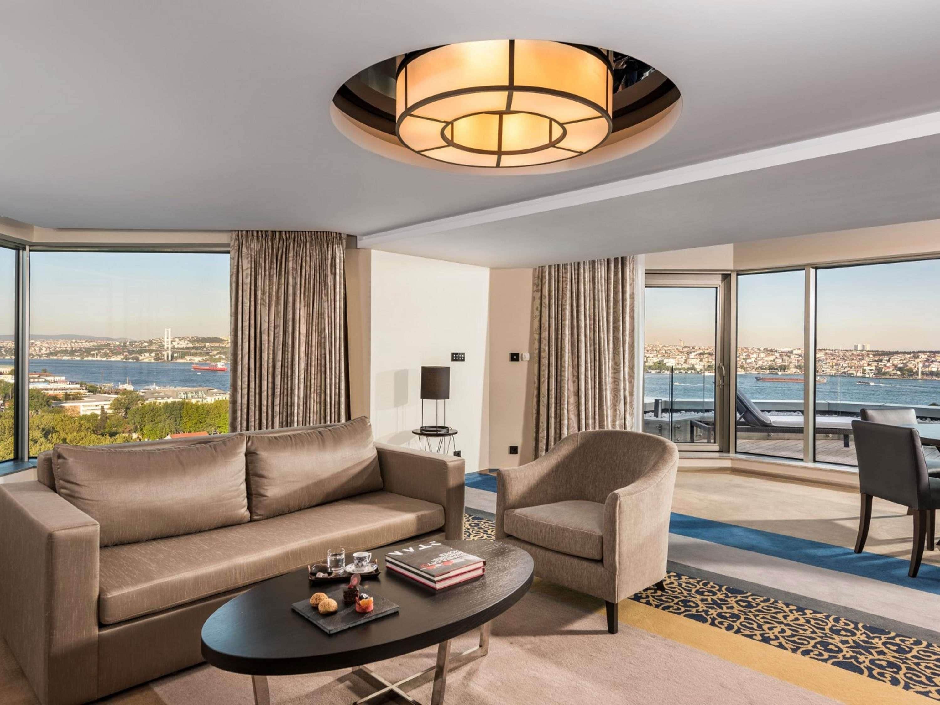 Una habitación de hotel moderna con sofá, sillón y una mesita con un libro y aperitivos.