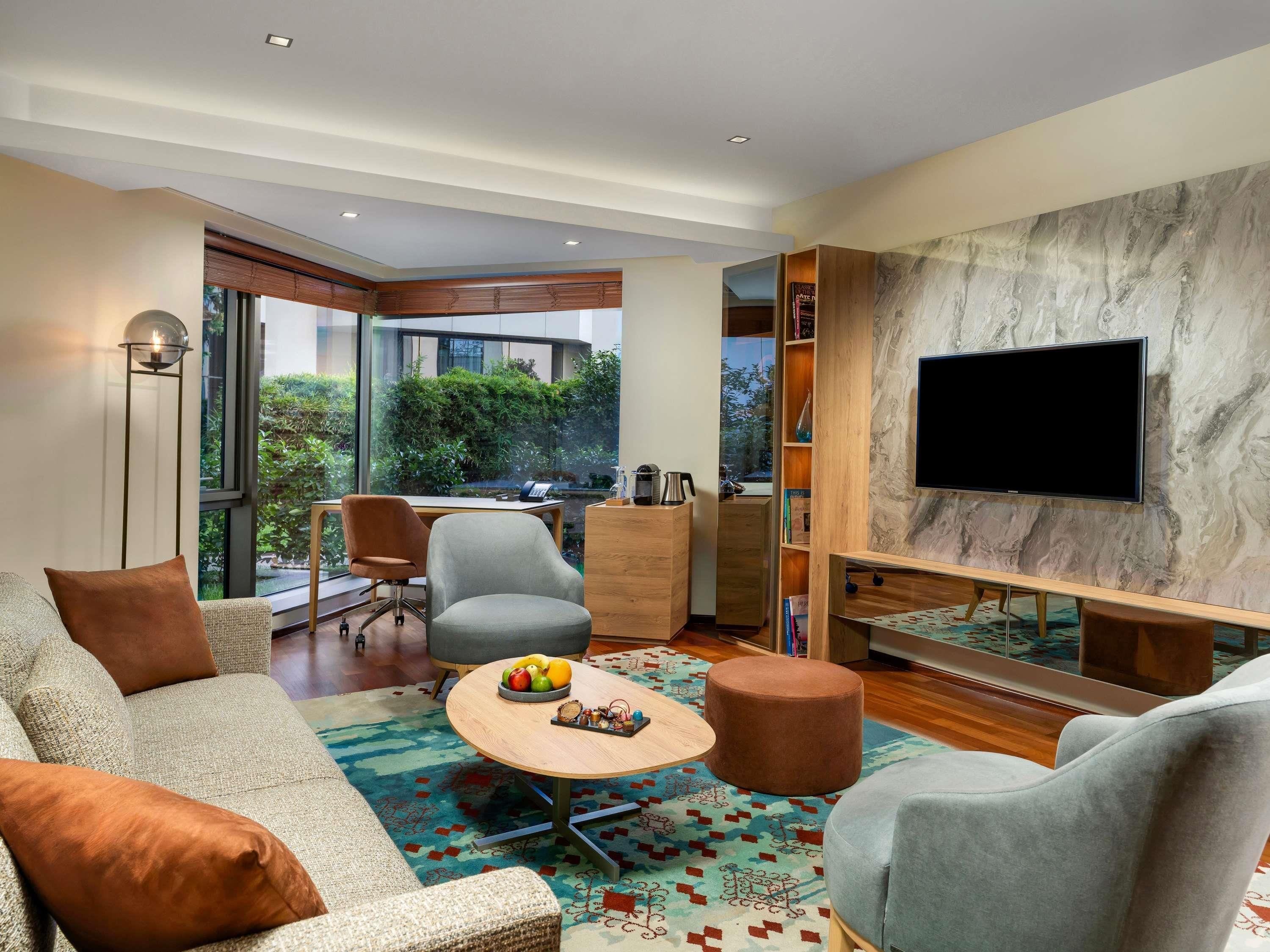 A modern living room with a sofa, armchairs, a coffee table with fruit, a TV on a marble wall, and a view of greenery outside.