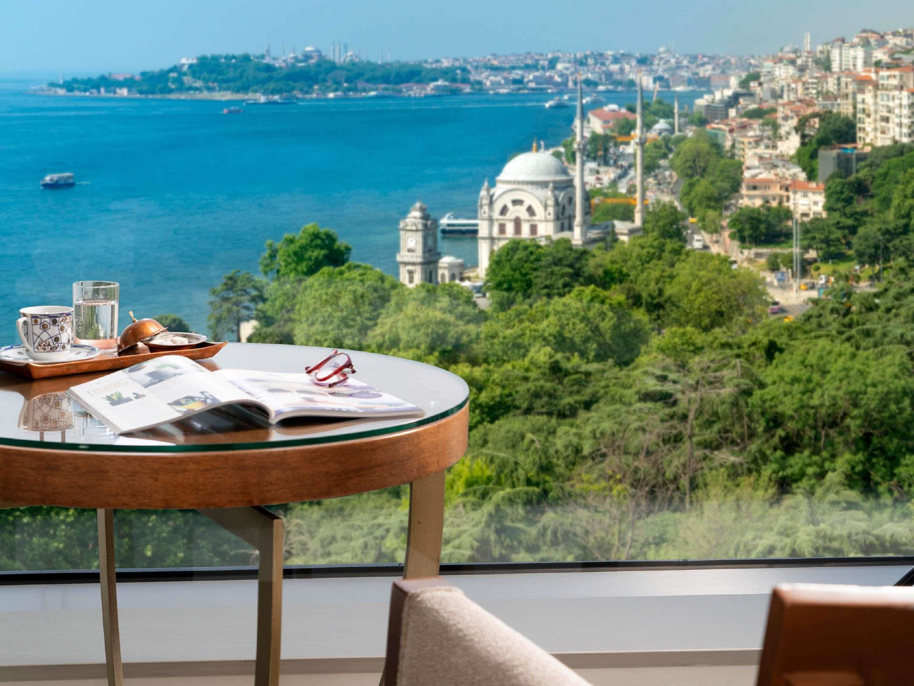 Una mesa con una bandeja de alimentos para el desayuno, un vaso de agua y una revista, con vistas a un paisaje urbano que incluye una masa de agua y un edificio destacado con una cúpula.