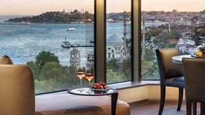 A room with a large window offering a view of a cityscape and a body of water. There are two chairs and a small table with a glass of wine and fruit. - Swissotel The Bosphorus Istanbul (Istanbul)